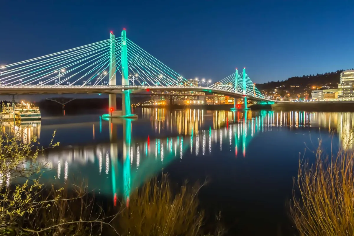 A nighttime view of a bridge reflected on calm water, with twinkling city lights in the background, evoking the lively yet serene atmosphere of Portland's waterfront.