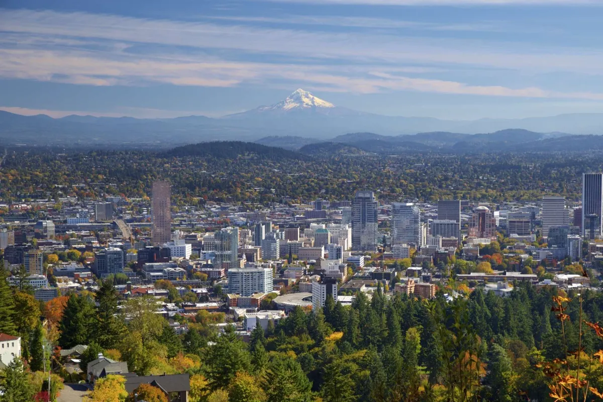 A sweeping view that captures Mount Hood in the distance, a city skyline, vibrant autumn foliage, and nearby mountains. The foreground features walking trails, hinting at nearby parks and outdoor adventures, while the overall mood is tranquil and inspiring—perfectly evoking the region's natural beauty.