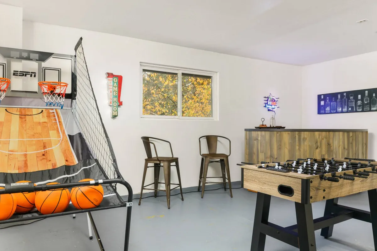 A fun, inviting recreation room with a basketball arcade game, foosball table, a bar area, and a large window with an autumn view. The space is designed for group activities and extra sleeping flexibility, especially for younger guests or night owls.