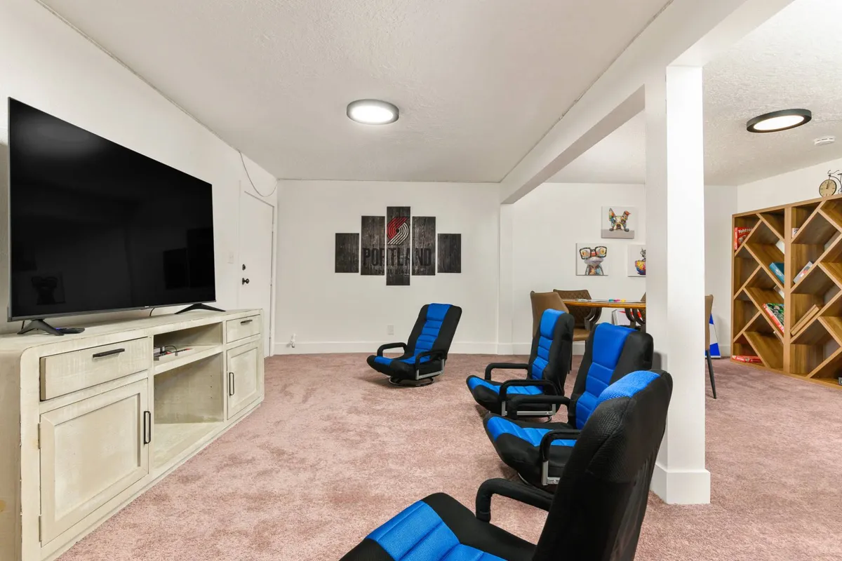 A dedicated gaming room featuring blue gaming chairs, a large television, decorative wall art, and a dining table. The room looks modern and purpose-built for immersive video gaming or casual hangouts, with cozy seating for multiple people.